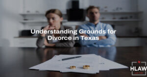 Divorcing couple sitting apart at a table with wedding rings and legal documents, illustrating no-fault divorce grounds in Texas, with text overlay "Understanding Grounds for Divorce in Texas" and HLAW logo.