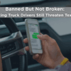 Truck driver holding a smartphone and texting while driving, illustrating the dangers of distracted commercial driving on Texas roads.